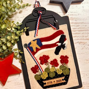 May include: A rectangular fabric wall hanging mounted on a black metal clip board. The fabric features an embroidered American flag with a gold star, a black eagle, and red geraniums in a black pot. A label on the pot reads 1776 star 2026.