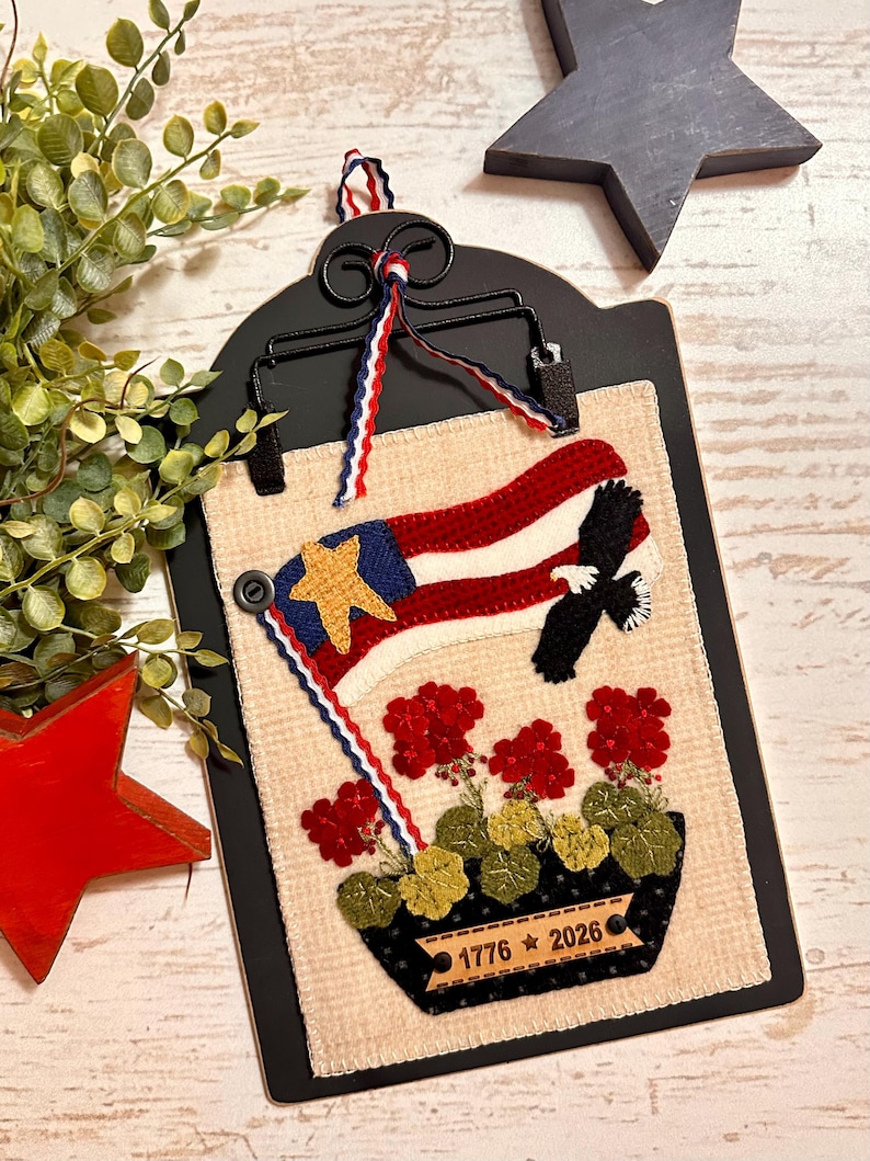 May include: A rectangular fabric wall hanging mounted on a black metal clip board. The fabric features an embroidered American flag with a gold star, a black eagle, and red geraniums in a black pot. A label on the pot reads 1776 star 2026.