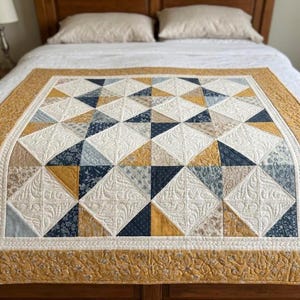 May include: A rectangular quilt featuring a geometric pattern of triangles in mustard yellow, navy blue, and cream. The cream sections have intricate feather-like stitching, while the border is a solid mustard yellow with a floral pattern.
