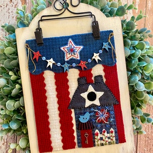 May include: A patriotic felt wall hanging with a red, white, and blue color scheme. The hanging features a house with a star and a bird, a string of stars and beads, and a red, white, and blue banner.
