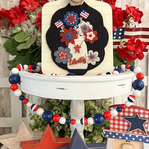 May include: A tiered tray decorated for the Fourth of July with a red, white, and blue beaded garland, a wooden cutout of a person wearing a red, white, and blue hat, and a wooden sign with a felt appliqu&eacute; of a mason jar filled with red, white, and blue flowers. The sign reads "Liberty".