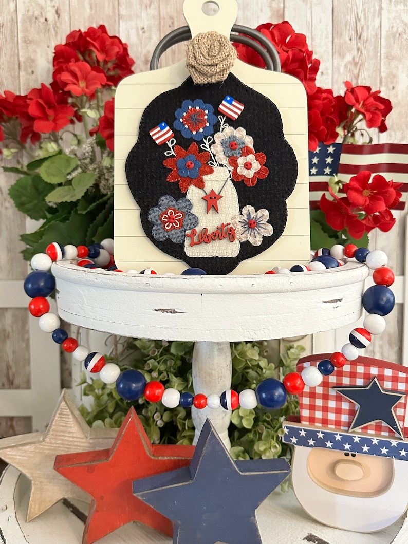 May include: A tiered tray decorated for the Fourth of July with a red, white, and blue beaded garland, a wooden cutout of a person wearing a red, white, and blue hat, and a wooden sign with a felt appliqu&eacute; of a mason jar filled with red, white, and blue flowers. The sign reads "Liberty".