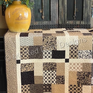 Op de afbeelding: Een patchwork quilt met een raster van vierkanten in tinten bruin, beige, cr&egrave;me en zwart met diverse bloem- en geometrische patronen. De quilt heeft cr&egrave;mekleurige tussenstroken en een rand met een klein bruin bloemenpatroon.
