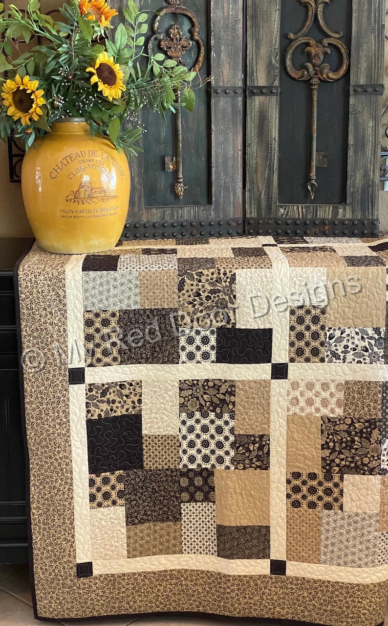 Op de afbeelding: Een patchwork quilt met een raster van vierkanten in tinten bruin, beige, cr&egrave;me en zwart met diverse bloem- en geometrische patronen. De quilt heeft cr&egrave;mekleurige tussenstroken en een rand met een klein bruin bloemenpatroon.