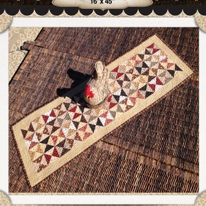May include: A rectangular table runner titled Heritage Runner measuring 16 inches by 45 inches. The runner features a patchwork design of small triangles in shades of red, cream, brown, and black, with a decorative fabric chicken placed on top.