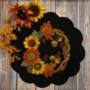 Puede incluir: Una corona de fieltro negro con temática otoñal. La corona está decorada con hojas, bellotas y flores de fieltro en tonos marrones, amarillos y naranjas.