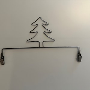 May include: A black metal Christmas tree-shaped wire clip holder. The tree is at the top, with a horizontal bar and two clips below. The clips are designed to hold photos or notes.