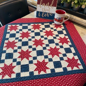 May include: A square quilt featuring a red, white, and blue star pattern with a red floral border. A white ceramic mug with a red truck graphic and a wooden sign with the text land that i love are placed on the quilt surface.