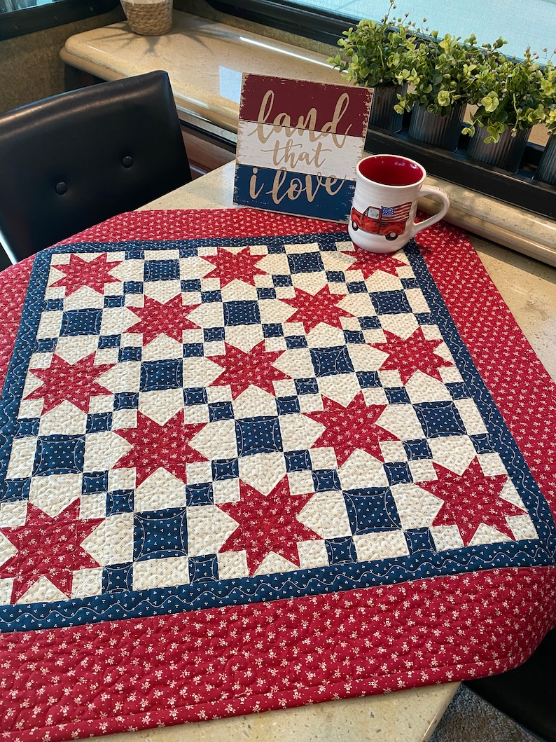 May include: A square quilt featuring a red, white, and blue star pattern with a red floral border. A white ceramic mug with a red truck graphic and a wooden sign with the text land that i love are placed on the quilt surface.