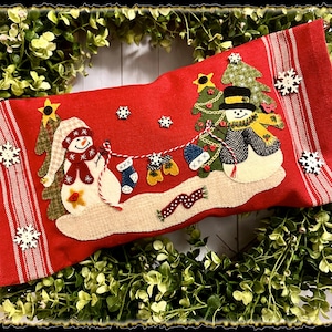 May include: Red and white striped stocking with a snowman and Christmas tree applique. The snowman is wearing a red scarf and a black hat. The Christmas tree is decorated with ornaments and lights. The stocking is decorated with snowflakes.