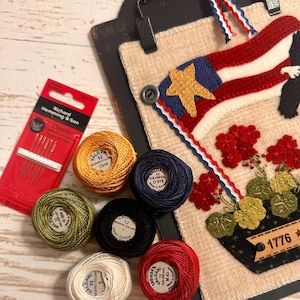 May include: A needlework project on a black clipboard featuring a flag, red flowers, and the text 1776, accompanied by six spools of Valdani pearl cotton thread in gold, navy, green, black, cream, and red, and a pack of Richard Hemming & Son needles.