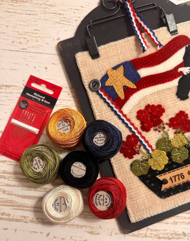 May include: A needlework project on a black clipboard featuring a flag, red flowers, and the text 1776, accompanied by six spools of Valdani pearl cotton thread in gold, navy, green, black, cream, and red, and a pack of Richard Hemming & Son needles.