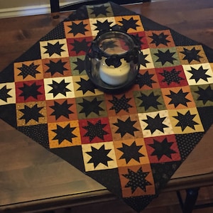 以下が含まれることがあります： 黒と茶色のパッチワークキルトで、星形の模様が施されています。キルトは木製のテーブルの上にあり、中央にはガラスの瓶に入ったろうそくがあります。