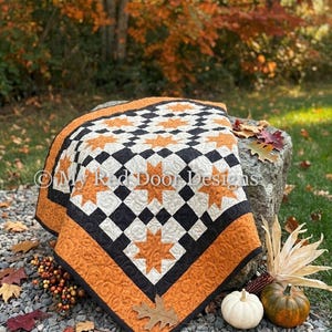 May include: A quilt featuring a repeating pattern of orange star shapes on a cream background with black square accents. The quilt has a wide orange border with a swirling quilted texture and the text My Red Door Designs printed on it.