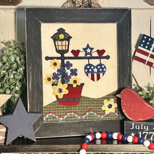 May include: A framed patriotic-themed wall art piece with a red, white, and blue heart, a lamppost with flowers, and two red birds. The frame is black with a distressed finish. The art piece is surrounded by red, white, and blue decorations, including a wooden star, a wooden bird, and a string of beads.