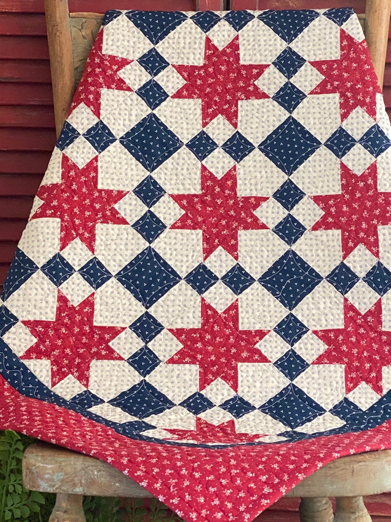 May include: A quilted blanket featuring a repeating star pattern in red, white, and navy blue. The fabric displays small floral prints throughout the design, with red stars, navy blue diamond shapes, and off-white square blocks stitched together.