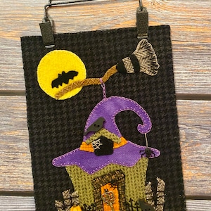May include: A black felt Halloween wall hanging featuring a purple witch's hat house, a yellow moon, a black bat, a broom, and a black cat.