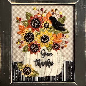 May include: Framed artwork with a pumpkin shape and the words "Give Thanks." It features felt sunflowers, leaves, and a black bird. The background is a grey and white checkered pattern, and the frame is black.