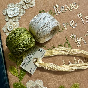 May include: Close-up of embroidery floss spools in green, white, and cream colors. The spools are labeled "Weeks Dye Works Pearl Collection" and "Lemonade". The floss is arranged on a brown fabric background with embroidered flowers and the text "Believe in your dreams".