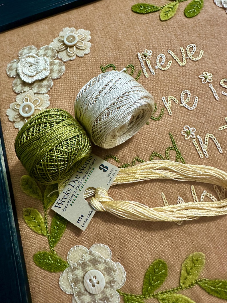 May include: Close-up of embroidery floss spools in green, white, and cream colors. The spools are labeled "Weeks Dye Works Pearl Collection" and "Lemonade". The floss is arranged on a brown fabric background with embroidered flowers and the text "Believe in your dreams".
