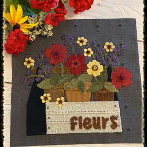 May include: A felt wall hanging with a rustic, farmhouse style featuring a wooden box with the word "fleurs" on it. The box is filled with potted red, yellow, and purple flowers.