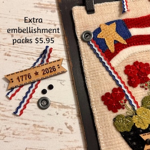 May include: A beige felt wall hanging featuring a red, white, and blue flag design with a gold star and red flowers. Included are a wooden tag engraved with 1776 and 2026, two small black buttons, one larger grey button, and red, white, and blue trim.