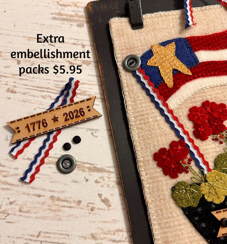 May include: A beige felt wall hanging featuring a red, white, and blue flag design with a gold star and red flowers. Included are a wooden tag engraved with 1776 and 2026, two small black buttons, one larger grey button, and red, white, and blue trim.