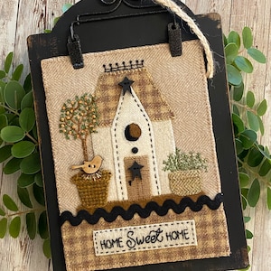 May include: A rustic wooden sign with a fabric birdhouse applique, a tree, and potted plants. The sign has a black frame and a jute rope for hanging. The words "HOME SWEET HOME" are stitched on the bottom, perfect for home decor.