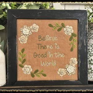 May include: A framed embroidery with a beige background and green floral accents. The embroidery reads "Believe There is Good in the World".