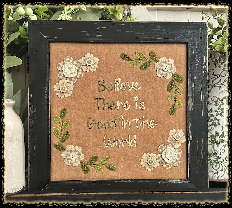 May include: A framed embroidery with a beige background and green floral accents. The embroidery reads "Believe There is Good in the World".