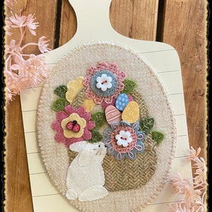 Könnte beinhalten: Ein weißer Filzhase schaut aus einem Korb voller bunter Filz-Ostereier und -Blumen. Der Korb ist von grünen Filzblättern umgeben und steht auf einem weißen Holzbrett.