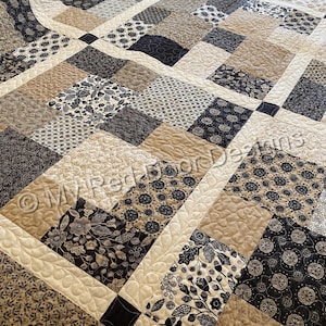 Op de afbeelding: Een patchwork quilt met een herhalend patroon van vierkante blokken in tinten zwart, cr&egrave;me en beige. De stoffen vierkanten tonen diverse bloemen-, blad- en geometrische prints, verbonden door cr&egrave;mekleurige tussenstroken met een doorgestikte textuur.