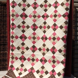 May include: A quilt featuring a repeating pattern of white, pink, and brown squares. The white sections have a quilted leaf texture. The bottom border is a dark brown fabric with a floral print in pink and white. The overall design is geometric.