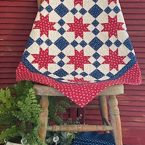 May include: A patchwork quilt featuring a repeating star pattern in red, white, and blue. The design incorporates red floral fabric, blue fabric with white dots, and cream fabric with small patterns. The quilt is draped over a wooden chair.