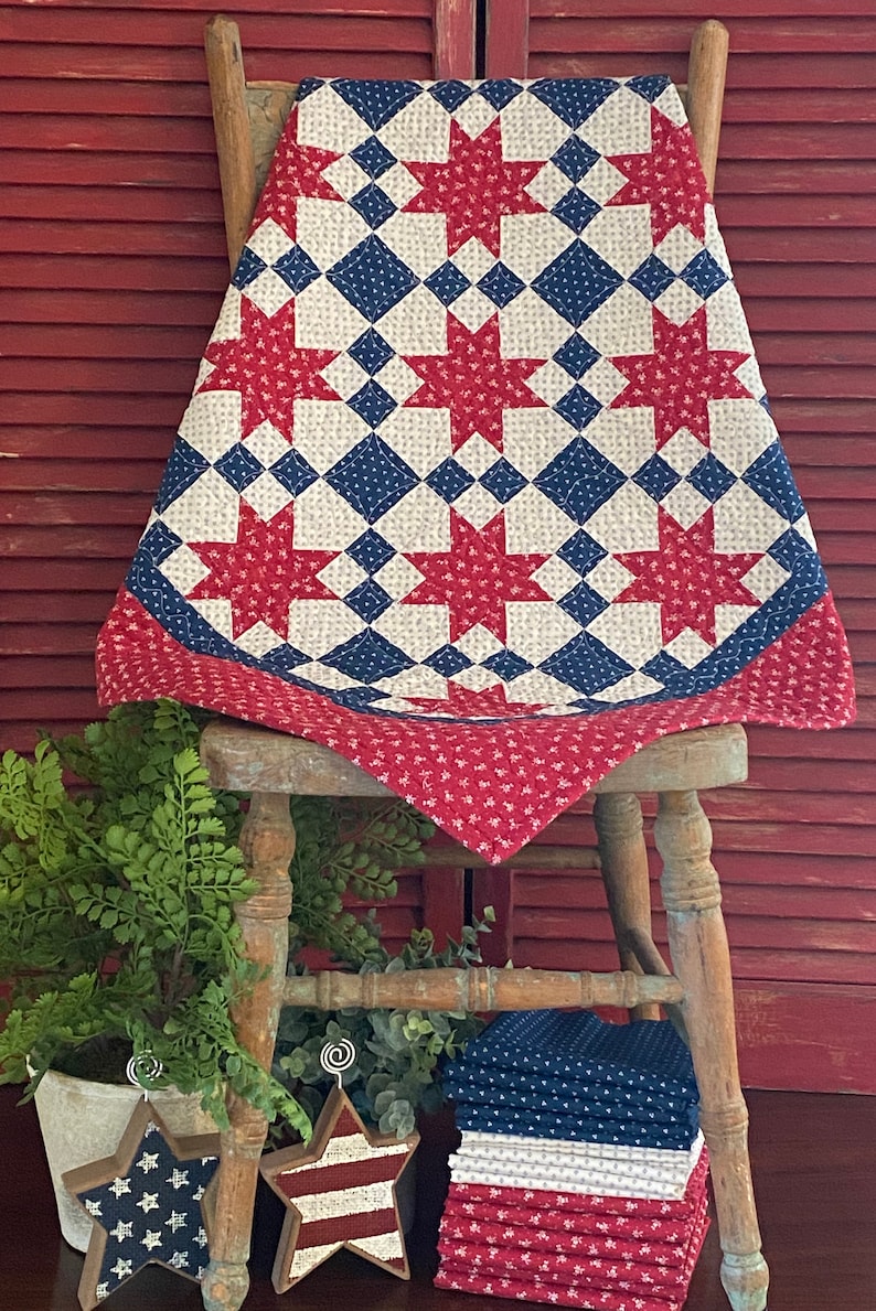 May include: A patchwork quilt featuring a repeating star pattern in red, white, and blue. The design incorporates red floral fabric, blue fabric with white dots, and cream fabric with small patterns. The quilt is draped over a wooden chair.