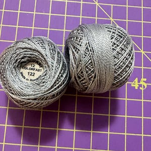 May include: Two silver-gray thread balls sit on a purple grid surface. One ball has a label that reads "12 COLORFAST 122 Made in Romania." The thread is tightly wound, showing the texture of the material. The image is well-lit, highlighting the thread's sheen.