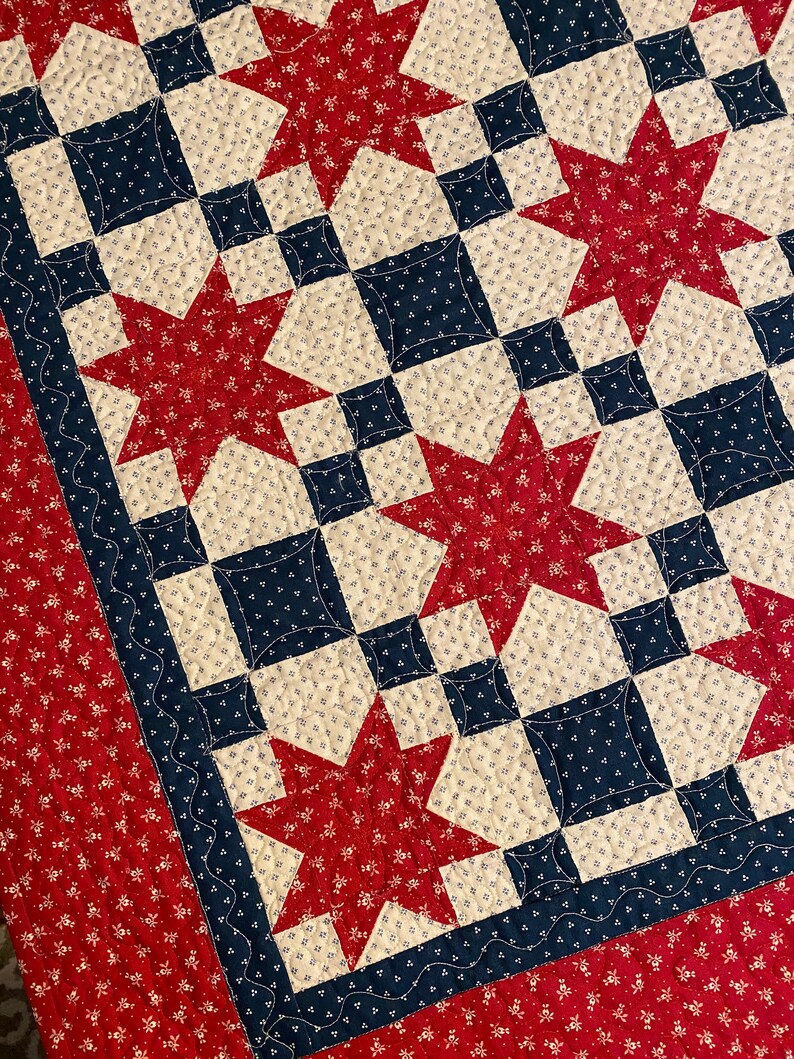 May include: A quilt featuring a repeating star pattern in red, navy blue, and cream. The fabric displays small floral and geometric prints. The design includes a wide red border with a floral pattern and a navy blue border with a wavy line stitch.
