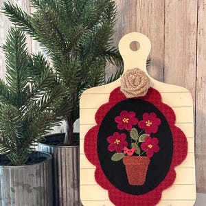 May include: A decorative wooden cutting board with a floral design. The board features a red scalloped border around a black oval with a felt flower pot and red flowers. A small burlap flower and a small bird are also present.