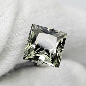 May include: A square-cut, pale green gemstone with a faceted surface that reflects light. The stone features sharp, pointed corners and a clear, translucent appearance, showcasing a geometric arrangement of triangular and trapezoidal facets.
