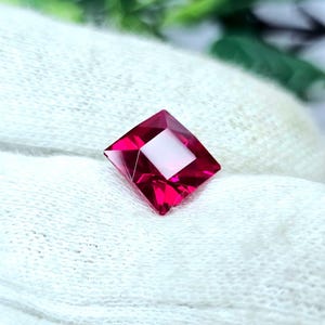 May include: A square-cut, vibrant red gemstone with faceted surfaces that reflect light. The stone features sharp, clean edges and a deep, saturated crimson color, showcasing a high level of clarity and a polished, brilliant finish throughout.