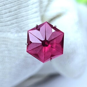 May include: A hexagonal, deep magenta gemstone featuring a faceted, star-like pattern on its surface. The stone is secured by small, silver-toned metal prongs at its edges, creating a precise, geometric appearance with sharp, clean lines.