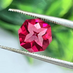 May include: A vibrant, deep red gemstone with a faceted, octagonal shape and a distinct four-pointed star pattern carved into its center. The gemstone is held securely by a pair of silver-toned metal tweezers, showcasing its reflective surface.
