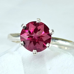 May include: A silver ring featuring a round, faceted, deep pink gemstone set in a six-prong setting. The stone displays a star-like pattern within its facets, and the band is a smooth, polished silver metal that tapers toward the setting.