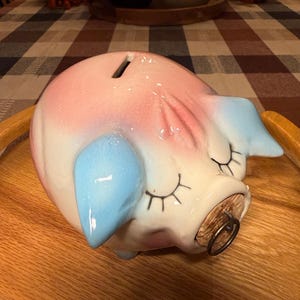 May include: A ceramic piggy bank featuring a glossy white finish with soft pink shading on the top and light blue accents on the ears. The pig has closed eyes with painted black eyelashes and a cork stopper in the snout with a metal ring pull.