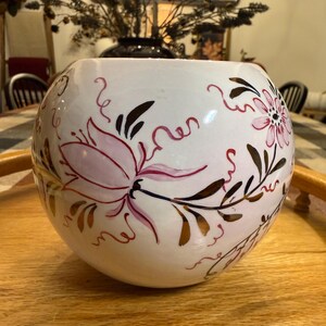 Puede incluir: Un jarrón esférico de cerámica blanca con motivos florales pintados a mano en tonos rosa y marrón oscuro. El diseño incluye flores estilizadas, delicadas enredaderas y hojas alargadas que envuelven el cuerpo redondeado del recipiente.