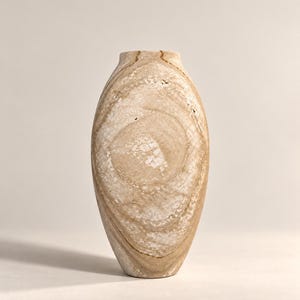 Könnte beinhalten: Eine hohe, oval geformte Vase aus Stein mit einem natürlichen, wirbelnden Muster in Beige-, Braun- und Weißtönen. Die Oberfläche des Gefäßes weist eine poröse, strukturierte Beschaffenheit mit sichtbaren organischen Maserungen und einer schmalen Öffnung auf.