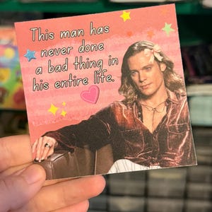 May include: A square sticker with a pink background featuring a person with long hair wearing a pink bow and a dark shirt. The text reads, This man has never done a bad thing in his entire life. The design includes a blue star, yellow stars, and a pink heart.