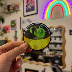 May include: A round sticker featuring a cartoon green alien with large eyes and antennae. The alien is set against a yellow and black background with a grid pattern. The sticker is held in a hand.
