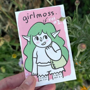 May include: A small, rectangular card with a pink border and the text "girlmoss." and "vol #1." features a cartoon elf with green hair and a green bag. The elf is wearing a white top and shorts. The card is held in a hand.