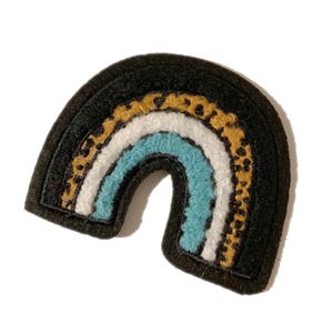 May include: A black felt rainbow patch with a leopard print section. The rainbow features stripes of white, light blue, and gold leopard print. The patch is a fun accessory for clothing or bags.
